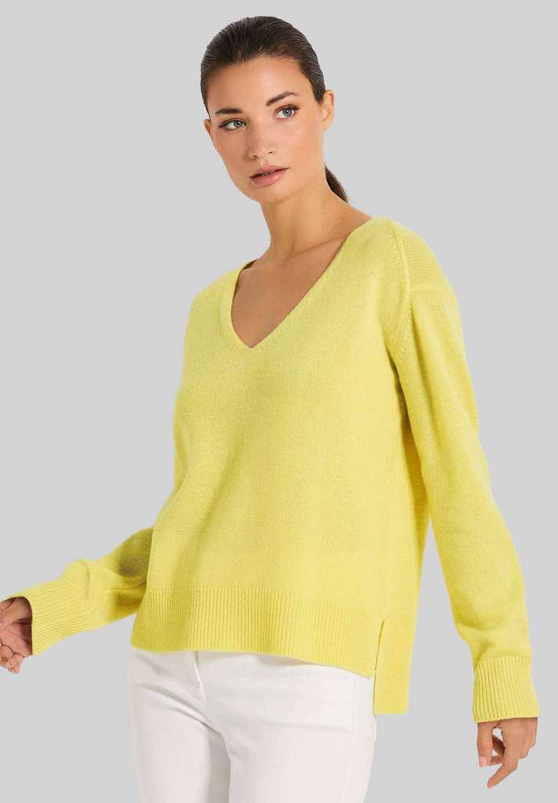 Gelbes V-Ausschnitt-Pullover aus weichem Material mit langen Ärmeln, geripptem Saum und seitlichen Schlitzen. Kombiniert mit weißen Hosen.