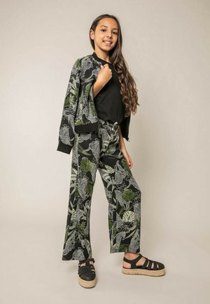 Jeune femme portant un haut noir, une veste et un pantalon assortis avec un imprimé feuilles vertes et léopard blanc, ainsi que des sandales compensées noires, debout devant un fond uni.