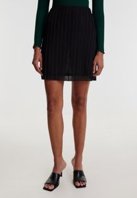 Schwarzer, plissierter Rock mit einem ausgestellten Design, der einen gerafften Taillenteil aufweist. Das Model trägt offen-toe schwarze Absatzpantoletten. Glatte Textur.