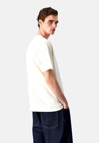 Cremfarbener T-Shirt mit kurzen Ärmeln und lässiger Passform, kombiniert mit dunklen Jeans. Glatte Stofftextur, einfaches Design ohne Muster oder Akzente.