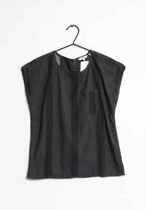 Blusa nera a maniche corte con strisce verticali di pizzo e tasca frontale, appesa su una gruccia nera su sfondo bianco.