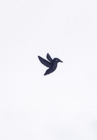 Tejido blanco con un logo de colibrí bordado en azul marino. La textura es suave con un tejido fino. Diseño minimalista y pequeño tamaño.