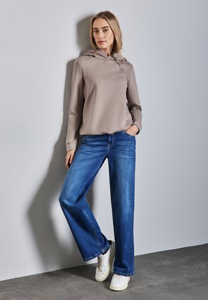 Hellbrauner Hoodie mit Kängurutasche, kombiniert mit weit geschnittenen blauen Jeans und weißen Turnschuhen, vor einer grauen Wand.