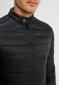 Zwart pufferjack met een staande kraag, verticale gestikte textuur, ritssluiting aan de voorkant en drukknoopsluiting details op de kraag. Zachte afwerking.