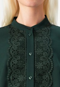 Tumši zaļa blouse ar augstu apkakli, sarežģītu mežģīņu detaļu gar priekšpusi un sešām dekoratīvām pogām. Gluda auduma tekstūra.