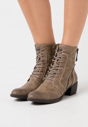 Bottes en daim taupe à hauteur de cheville avec lacets sur le devant, fermetures éclair sur les côtés, couture blanche et talons bas sur des jambes nues contre un fond blanc.