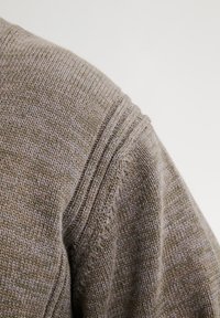 Manica di un maglione di lana lavorato a maglia marrone che mostra in dettaglio la cucitura a coste sulla spalla, su uno sfondo uniforme.