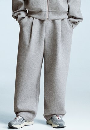 Osoba oblečená v volných šedých teplákách, s rukama v kapsách a v šedých sportovních teniskách, stojící na světlem pozadí.