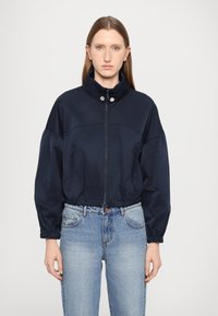 Beskjært marineblå jakke med høy krage, glidelås foran, elastiske mansjetter og to sidelommer, kombinert med lyseblå jeans.