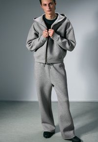 Harmaa huppari ja siihen sopivat collegehousut pehmeästä puuvillasekoitekankaasta. Hupparissa on vetoketju edessä ja suuret taskut. Rennon istuvuuden omaava malli.