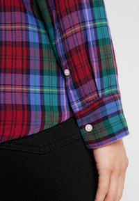 Gros plan sur le bras d'une personne portant une chemise à carreaux multicolores avec des boutons et un pantalon noir sur un fond clair uni.