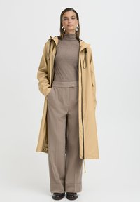 Gai beige garais lietusmētelis ar aukliņām, uzvilkts virs brūna turtlenecka un gaišbeigām platiem biksēm, ar tumšiem akcentiem un brīvu griezumu.
