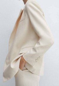 Blazer de crema ajustado y pantalones a juego, con la mano casualmente en el bolsillo, mostrando un atuendo empresarial minimalista y elegante.