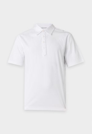 Personne debout portant un polo blanc à manches courtes, un jean bleu et des baskets marron, avec une main dans la poche sur fond blanc.