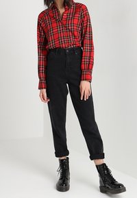 Chemise à carreaux rouges à manches longues, boutonnée, associée à un pantalon noir taille haute et des bottines noires à lacets.