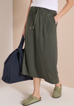 Femme portant une jupe midi vert olive, un haut blanc, des chaussures vertes sans lacets, tenant un grand sac bleu marine à côté d'elle.