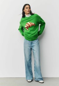 Zaļš adīts džemperis ar brūnu un bēšu kruasāna dizainu priekšpusē, kombinēts ar gaiši ziliem platām kājām šortiem un baltām sporta apaviem.