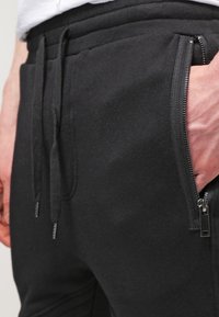 Černé joggingové kalhoty vyrobené z hladké látky, s páskem na stažení a boční kapsou na zip s kovovým hardwarovým prvkem.