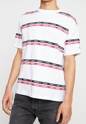 Vit bomulls t-shirt med röda och svarta horisontella ränder, försedd med upprepad text "TOMMY JEANS" och rundad halsringning.