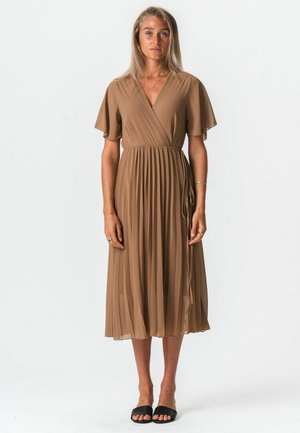 Žena stojí oblečená v midi opásaných opálených šatách s rozviatymi rukávmi a plisovanou sukňou, v kombinácii s čiernymi šlapkami s otvorenou špičkou, neutrálne pozadie.