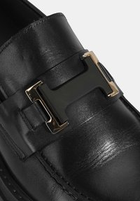 Schwarze Lederloafer mit glänzender Oberfläche, einem breiten Riemen mit goldfarbenem und schwarzem Hardware-Akzent sowie detaillierter Naht entlang der Kante.