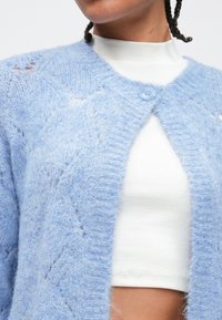 Ženska, oblečena v svetlo modro mehko jopico z enim gumbom, pripetim čez bel rebrast kratki top, ki kaže del njenega trebuha.