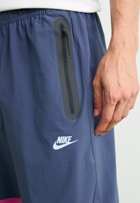 Gros plan sur une main à côté d'un short bleu Nike avec une poche noire verticale zippée et un logo blanc sur la cuisse.