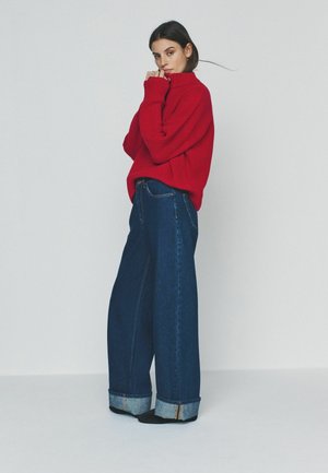 Kobieta w czerwonym swetrze z golfem i szerokich ciemnoniebieskich dżinsach stojąca na tle jednolitego, jasnego tła.