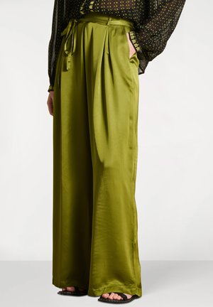 Pantalon large en satin vert avec plis et ceinture à la taille, porté avec un chemisier noir à motifs et des sandales noires à bout ouvert.