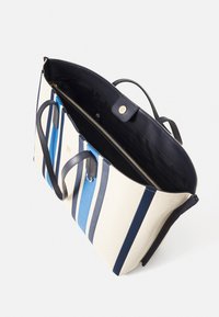 Bolso tote a rayas con rayas azules y marino sobre una lona color crema. Presenta asas de cuero negro y un compartimento interior con cremallera.