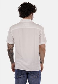 Camicia bianca a maniche corte con tessuto liscio, colletto classico e cuciture soft sulle spalle. Indossata con jeans blu casual.