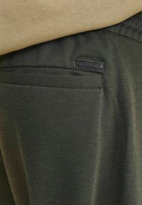 Tamnozelena tkanina izbliza, s glatkom teksturom i vidljivim stražnjim džepom. U kutu se nalazi crna oznaka "JACK & JONES".