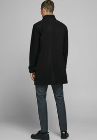 Melns vilnas mētelis ar piegriezumu, ar augstu apkakli. Valkāts kopā ar pelēkiem kostīmpantiem ar rūtiņām un melnām sporta apaviem, skatīts no aizmugures.