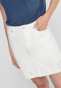 Jupe en denim blanc avec un ourlet effiloché, des détails déchirés et un design classique à cinq poches, avec un bouton en argent à la taille.