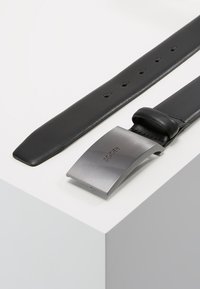 Cinturón de cuero negro con hebilla de metal plateado mate. La hebilla cuenta con un logo grabado y un diseño aerodinámico. Presenta varios agujeros de ajuste.