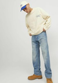 Beige sweatshirt with blue text, light blue jeans, and tan work boots. The model wears a white cap with a blue brim.