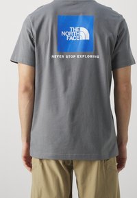 Harmaa puuvillapaita, jossa on sininen neliögraafikko, jossa lukee "THE NORTH FACE" -logo ja "NEVER STOP EXPLORING" -teksti valkoisella. Klassinen malli.
