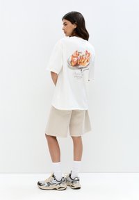 Vit oversized t-shirt med en färgglad efterrättsgrafik på ryggen, kombinerad med beige shorts och svarta-silvriga träningsskor.