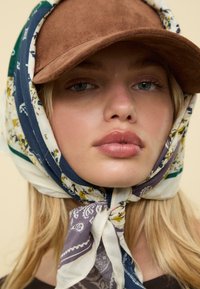 Casquette en daim marron portée avec une écharpe à motifs aux couleurs bleue, blanche et verte. Tissu noué autour du cou ; texture douce.