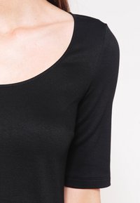 Top negro de manga corta con un escote amplio, textura suave y diseño ajustado, que resalta la zona de los hombros y la parte superior del cuerpo.