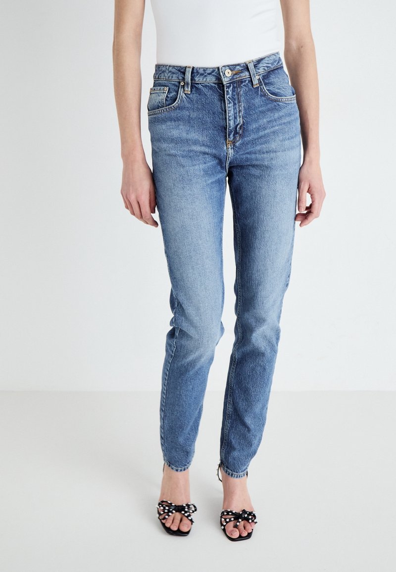 Femme portant un jean skinny taille haute bleu clair et des talons noirs ouverts avec des détails de nœud à pois, debout sur un fond uni.