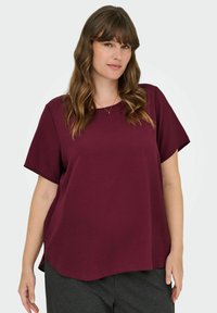 Burgundy kortärmad t-shirt med rundad halsringning och avslappnad passform, tillverkad av en blandning av bomull. Sömmen är något böjd.