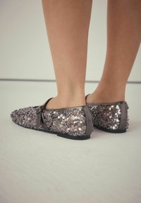 Flache Schuhe mit einer abgerundeten Zehenpartie, bedeckt mit glänzenden silbernen Pailletten und einem grauen Hintergrund. Sie verfügen über einen dünnen Riemen über dem Spann.