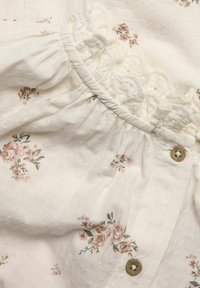 Hvidt stof med et tekstureret mønster, prydet med små rosa og grønne blomstermotiver. Flæset blondekant og to træknapper på siden.