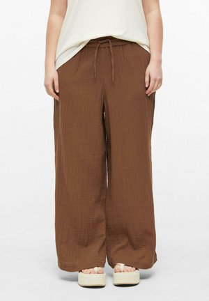Personne portant un pantalon marron ample avec un cordon à la taille et des sandales blanches à plateforme, debout devant un fond uni.