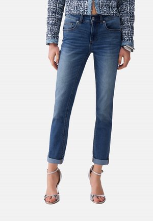 Donna con jeans blu aderenti risvoltati, maglia a maniche lunghe con fantasia e sandali argento con tacco alto, in piedi davanti a uno sfondo semplice.