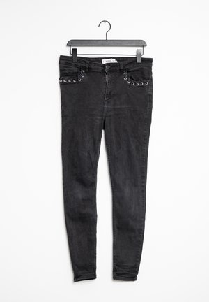 Jean noir en denim, avec une coupe ajustée, des œillets en métal argenté décoratifs le long des poches, et une fermeture éclair classique.