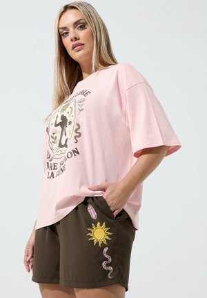 Lyshåret kvinde iført en oversized pink t-shirt med et himmelsk design og brune shorts dekoreret med sol-, krystal- og slangegrafik.
