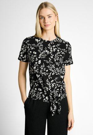 Blonde vrouw draagt een zwart bloemig kortemouwen shirt met strik aan de voorkant, staat met handen in de zakken en een neutrale uitdrukking tegen een witte achtergrond.