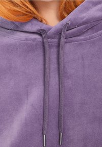 Sudadera de forro polar morada con dos cordones que presentan puntas de metal plateadas. Textura suave y corte holgado, con un bolsillo tipo canguro en la parte delantera.
